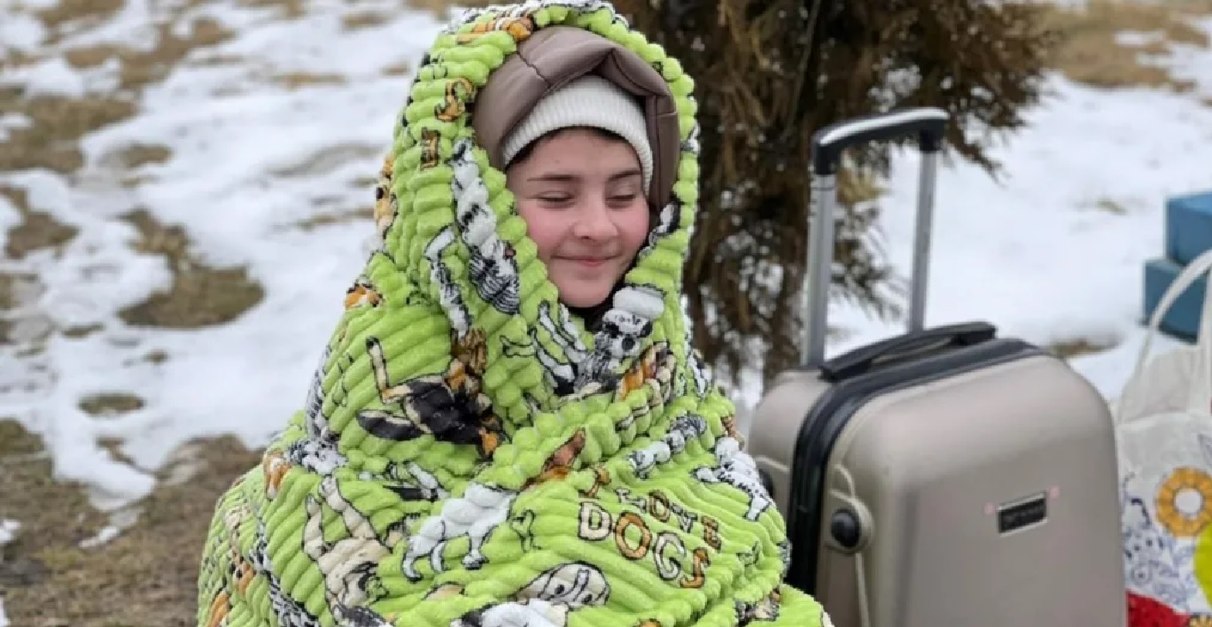 Vaikas Ukrainos pabėgėlis lauke su lagaminu, šildosi įsisupęs į Kikos komandos padovanotą medvilninį pledą