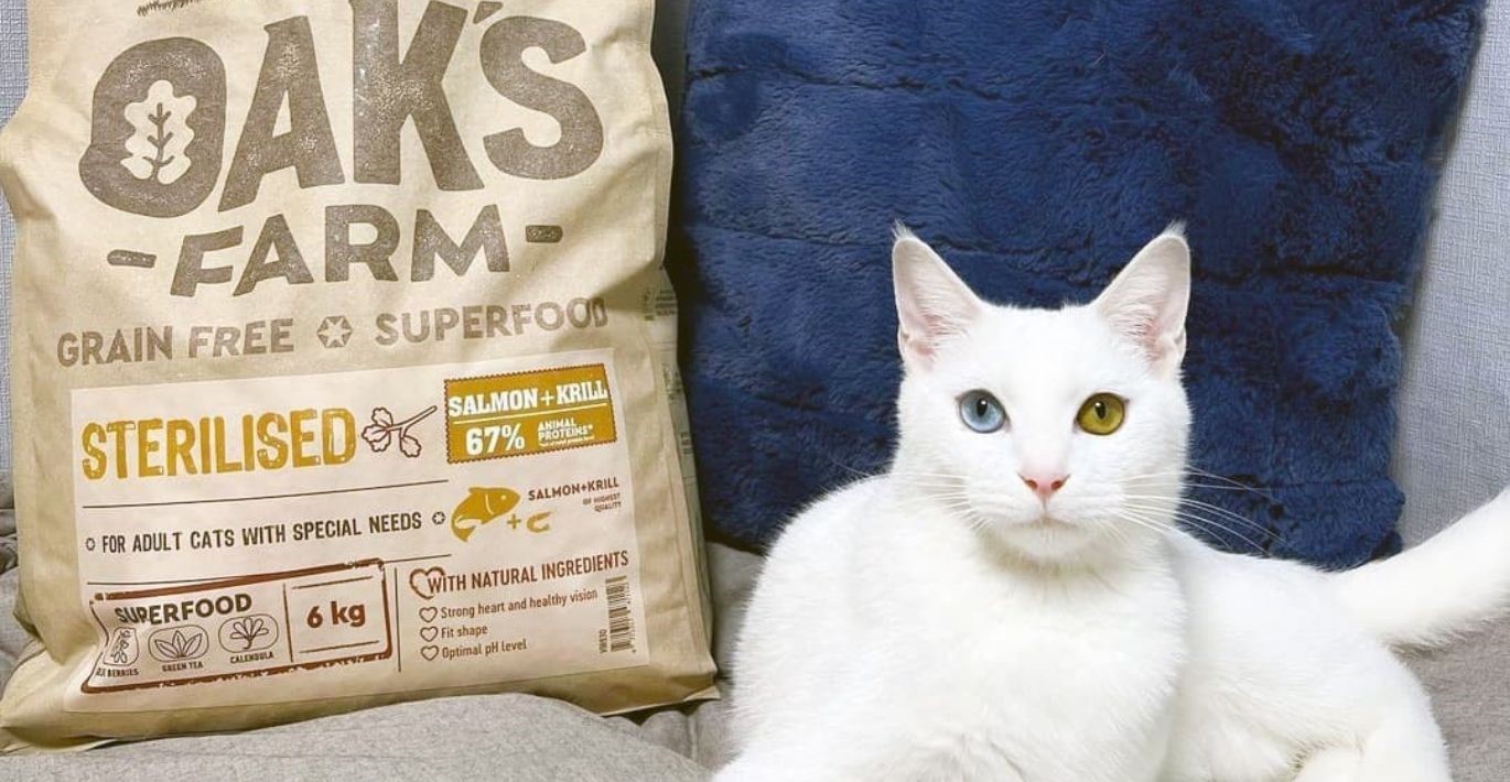 Baltakailis katinas su išraiškingomis akimis pozuoja šalia Oak‘s farm pašaro pakuotės